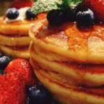 Pile de pancakes au yaourt moelleux, garnis de framboises, de myrtilles et d'un filet de miel, sur une assiette blanche.
