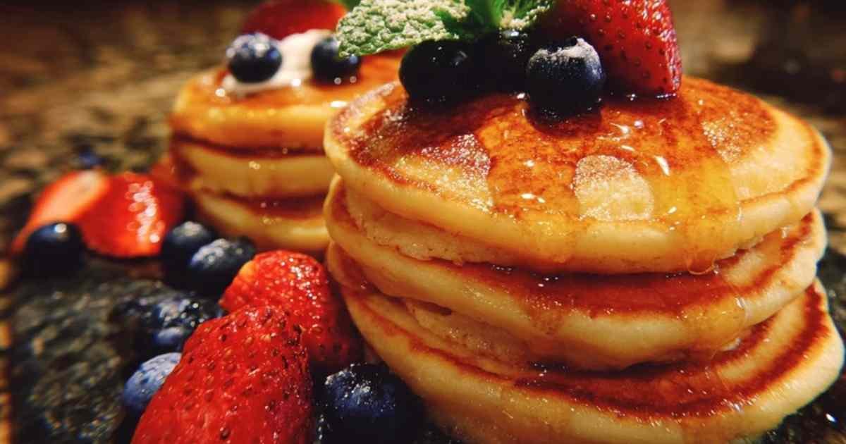 Pile de pancakes au yaourt moelleux, garnis de framboises, de myrtilles et d'un filet de miel, sur une assiette blanche.