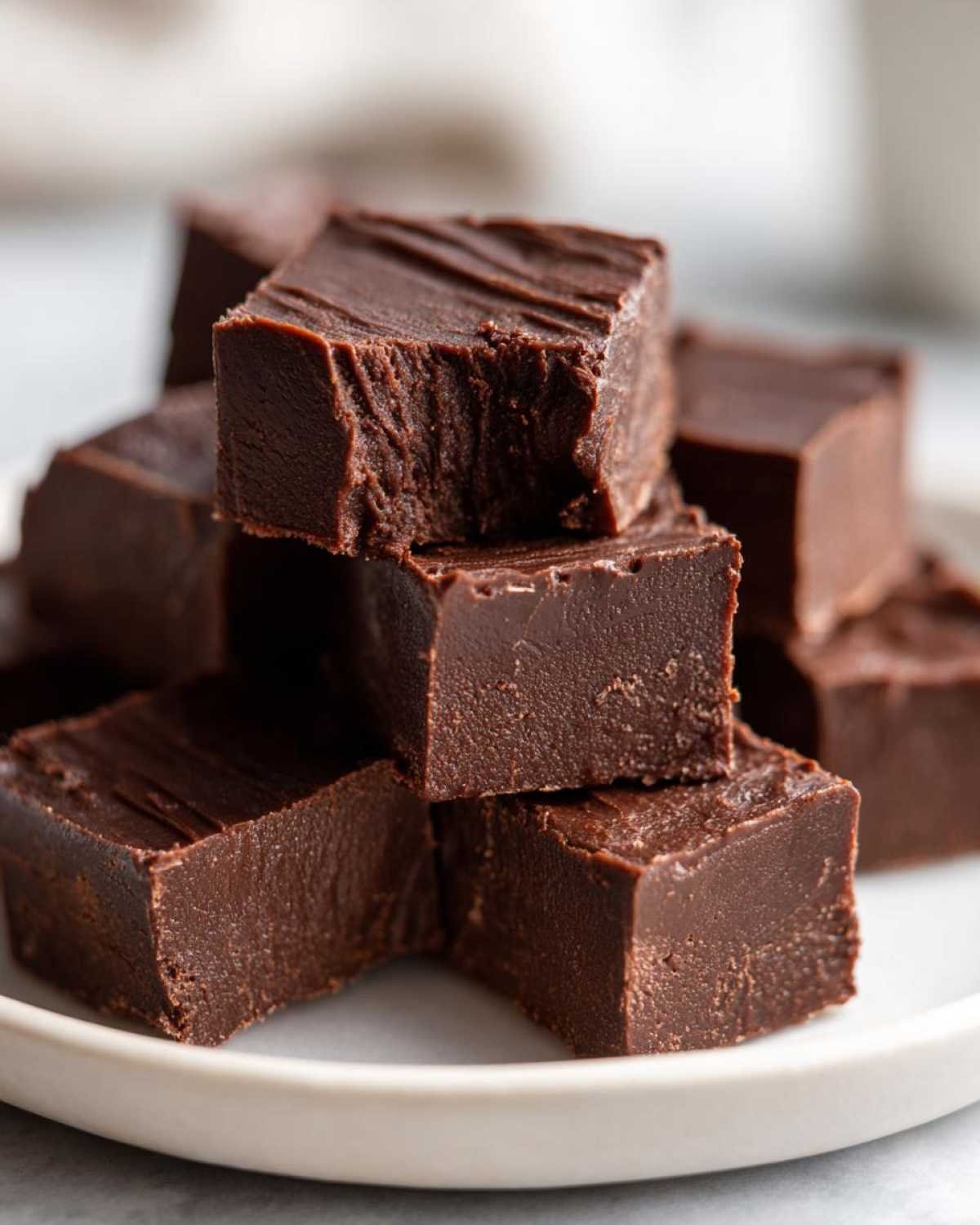 Gros plan de carrés de fudge keto 3 ingrédients au chocolat, empilés sur une assiette blanche.