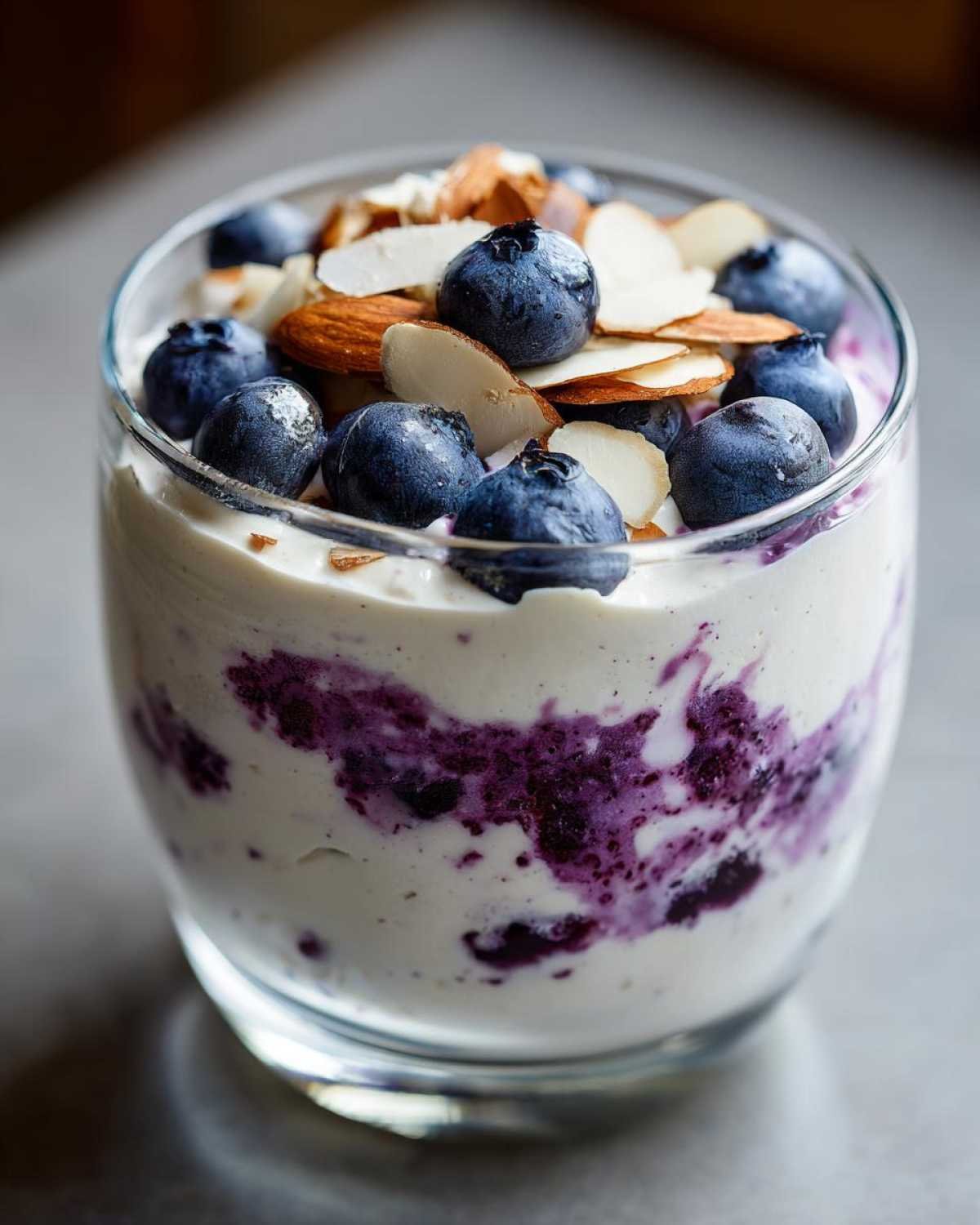 Gros plan d'une mousse cottage cheese myrtilles dans un verre, garnie de myrtilles fraîches et d'amandes effilées.