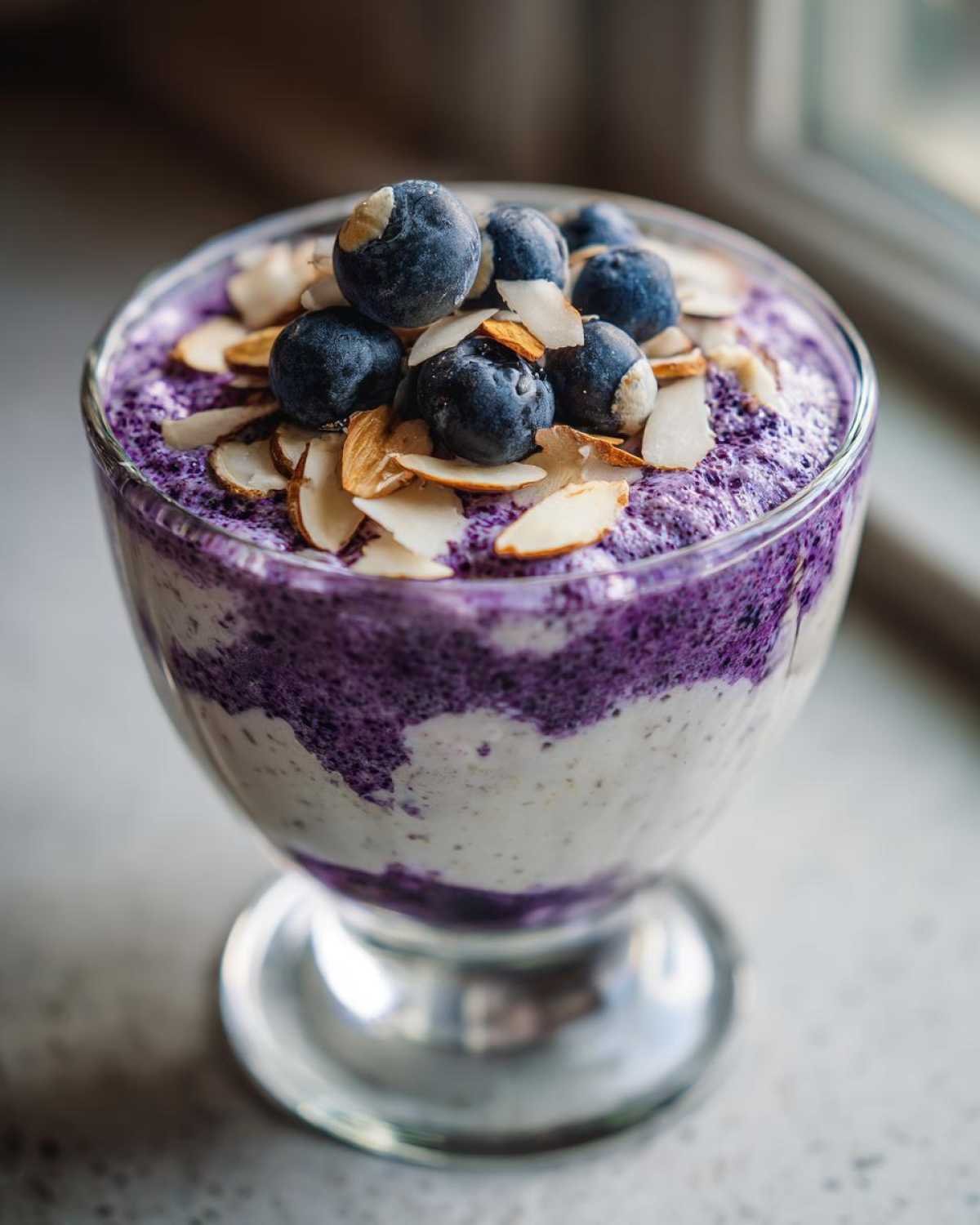 Gros plan d'une mousse cottage cheese myrtilles dans un verre, garnie de myrtilles fraîches et d'amandes effilées.