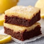 Gros plan de deux brownies au citron facile empilés, avec une base chocolatée foncée et un dessus crémeux jaune pâle, saupoudrés de sucre glace.