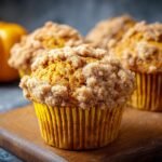Gros plan sur des muffins au potiron avec une garniture crumble sur une planche en bois, avec une citrouille en arrière-plan.