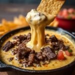 Une tortilla chip trempée dans une trempette Queso au Bœuf chaude et filante, garnie de morceaux de bœuf et de tomates.